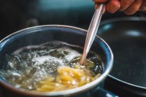 Fermeture de la récolte non reconnaissable cuisson à la main pâtes crues se répandant de la boîte à la casserole avec de l'eau bouillante pendant la préparation des aliments dans la cuisine — Photo de stock