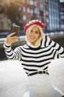 Joven mujer sonriente en gorra roja francesa, blusa a rayas y pantalones cortos blancos tomando fotos sobre fondo urbano - foto de stock