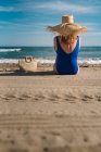 Вид ззаду красива жінка в капелюсі і купальник сидить з сумкою на піщаному узбережжі, дивлячись на хвилі під бірюзовим хмарним небом — стокове фото