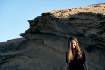 Joven mujer de pelo largo con estilo mirando hacia abajo mientras está de pie en la luz del sol con roca en el fondo - foto de stock