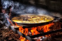 Grande casserole en fer avec bouillon bouillant pour cuisiner la paella sur un feu ouvert avec du bois — Photo de stock