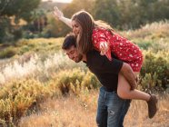 Fröhliches Paar, das Huckepack spielt, während es an einem Sommertag im gelben Gras der Landschaft steht — Stockfoto