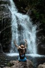 Веселая женщина-путешественница в шляпе улыбается и смотрит в камеру, сидя на мокром валуне возле водопада — стоковое фото