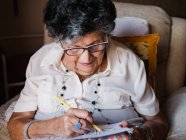 Mujer mayor en camisa blanca y gafas dibujo sobre papel con lápiz sentado en sillón en casa - foto de stock
