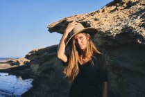 Joven mujer de pelo largo reflexiva mirando hacia la costa rocosa - foto de stock