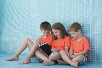 Діти в яскраво-помаранчевих футболках читають цікаву книгу, сидячи на дні порожнього басейну — стокове фото