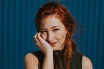 Attraktive junge Frau mit roten Haaren blickt in die Kamera als berührendes Gesicht am sonnigen, hellen Tag vor blauer Wand — Stockfoto