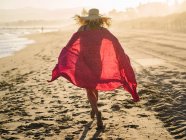 Невпізнавана жінка в капелюсі махає червоним шарфом і бігає уздовж узбережжя в сонячний яскравий день — стокове фото