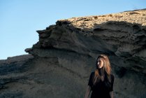 Молодая задумчивая женщина, стоящая на солнце с камнем на фоне — стоковое фото