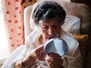 Seniorin trägt Lippenstift auf, während sie in den Spiegel schaut und zu Hause auf einem Sessel sitzt — Stockfoto