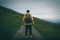 Back view of unrecognizable male with backpack standing on rough path on grassy hill slope against gray overcast sky in nature — Stock Photo