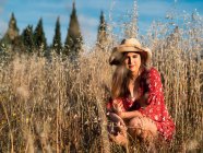 Attraente donna sorridente in cappello di paglia e vestito rosso seduta su campo selvaggio sullo sfondo del cielo blu — Foto stock