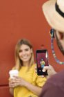 Jovem posando com café inclinado na parede e homem irreconhecível tirando foto com telefone celular — Fotografia de Stock