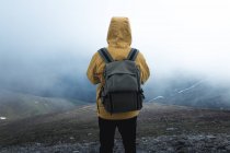 Vista trasera de un tío con mochila en la ladera de la colina contra la niebla gruesa durante el viaje en la naturaleza. - foto de stock