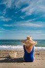 Vista posterior de la mujer bonita en sombrero y traje de baño sentado con bolsa en la playa de arena mirando las olas bajo el cielo turquesa nublado - foto de stock