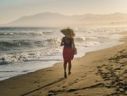 Vista posteriore di donna magra in cappello e pareo che corre lungo la costa in piena luce del sole — Foto stock