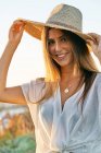 Jovem mulher em roupas brancas e chapéu de palha elegante sorrindo e olhando para a câmera na natureza — Fotografia de Stock