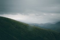 Вид сзади неузнаваемого самца с рюкзаком, идущего по неровной дорожке по склону холма на фоне пасмурного неба в природе — стоковое фото