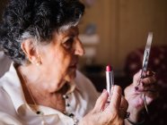 Seniorin trägt Lippenstift auf, während sie in den Spiegel schaut und zu Hause auf einem Sessel sitzt — Stockfoto