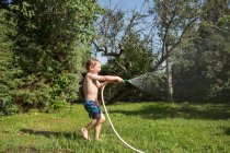Little laughing kid in shorts and with bare feet splashing water from garden hose — Stock Photo