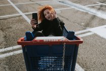 Smiling woman with smartphone in shopping trolley in parking lot — Stock Photo