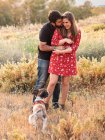 Jovem casal feliz abraçando e se divertindo com pouco cão entre grama alta no campo — Fotografia de Stock