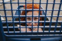 Attractive young woman with red hair sitting in blue shopping cart and looking in camera through trolley grate — Stock Photo