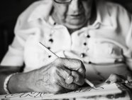 Anziana in camicia bianca e occhiali da disegno su carta con matita seduta su poltrona a casa — Foto stock