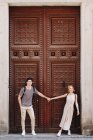Joven pareja alegre y juguetona en ropa casual tomados de la mano frente a la hermosa puerta vieja - foto de stock