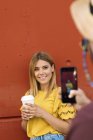 Mujer joven posando con café apoyado en la pared y hombre irreconocible tomando fotos con teléfono móvil - foto de stock