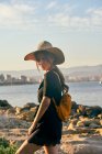 Молодая туристка в соломенной шляпе и рюкзаке, стоящая возле пляжа — стоковое фото