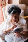 Mujer mayor que aplica lápiz labial mientras se mira en el espejo y se sienta en el sillón en casa - foto de stock