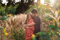 Nachdenklicher Mann umarmt lächelnde schwangere Frau im Hintergrund des malerischen grünen Parks bei sonnigem Tag — Stockfoto