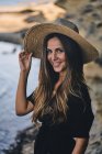 Молодая длинноволосая улыбающаяся женщина в шляпе смотрит в камеру на пляже — стоковое фото