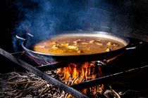 Grande padella di ferro con brodo bollente per cuocere la paella sul fuoco aperto con legna — Foto stock