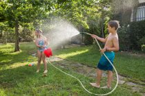 Junge in Badehose spritzt an einem sonnigen Tag Wasser aus Gartenschlauch auf ein Mädchen — Stockfoto