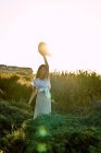Молода жінка в білому одязі дивиться на камеру і тримає капелюх в природі на заході сонця — стокове фото