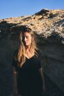 Young pensive woman looking down while standing in sunlight with rock on background — Stock Photo