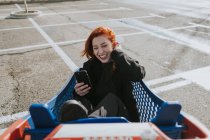 Mujer sonriente con teléfono inteligente en el carrito de la compra en el estacionamiento - foto de stock