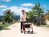 Felice donna anziana in camicetta con walker nel parco tropicale nella giornata di sole — Foto stock