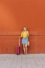 Молода жінка туристка з валізою розмовляє на мобільному телефоні, спираючись на червону стіну — стокове фото