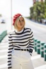 Young blonde woman in striped black and white shirt and red French cap walking on blurred city background — Stock Photo