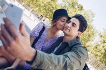 Joven hombre guapo tomando selfie con novia mientras se besa en hermoso jardín - foto de stock