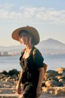 Joven turista con sombrero de paja y mochila de pie cerca de la playa - foto de stock