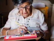 Senior femme en chemise blanche et lunettes dessin sur papier avec crayon, assis sur fauteuil à la maison — Photo de stock