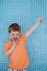 Joyful male kid in bright orange T-shirt showing gesture of rock on background of tiled wall of pool — Stock Photo