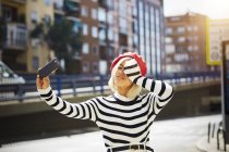 Giovane bella donna sorridente in berretto rosso francese, camicetta a righe e pantaloncini bianchi scattare foto su sfondo urbano — Foto stock