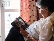 Зверху сіро волохата жінка в окулярах дивиться на фото онуки в альбомі вдома — стокове фото