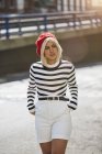 Молодая блондинка в полосатой черно-белой рубашке и красной французской кепке ходит на размытом фоне города — стоковое фото