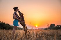 Mujer joven entrenando perro grande en la naturaleza salvaje en el fondo con el sol poniente naranja. Perro saltando alto para el placer - foto de stock